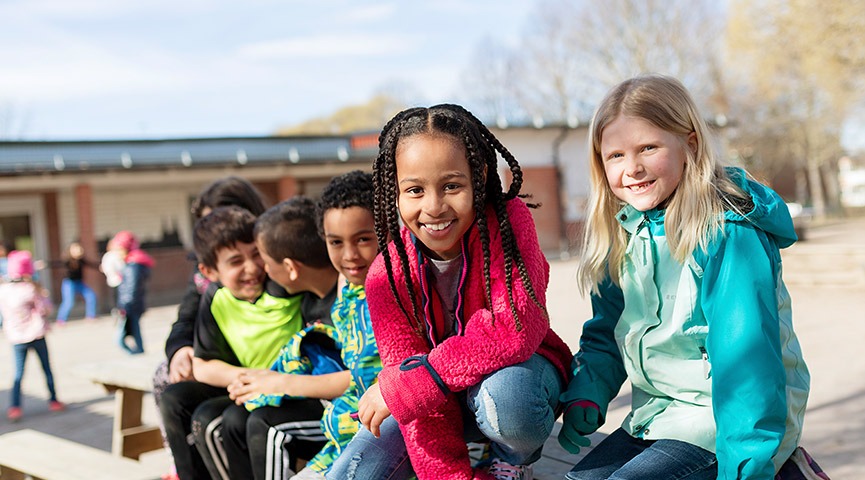 En grupp lågstadieelever sitter på en skolgård och tittar in i kameran