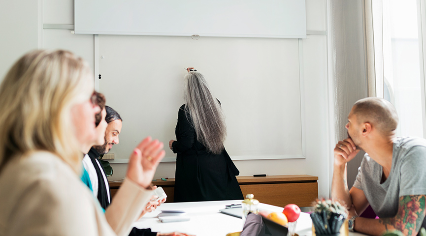 En person skriver på en whiteboard-tavla medan kollegor sitter runt ett bord